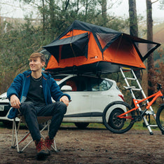 A mountain biker camping with his  TentBox Lite 1.0 Orange Edition on a white hatchback