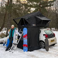 A woman with a snowboard smiling in front of her TentBox Lite car roof tent
