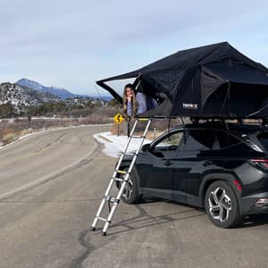 A happy woman enjoying the view from her TentBox Lite 1.0 which is installed on the roof of a black SUV