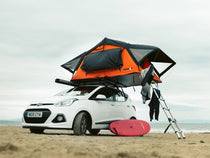 Small white car parked on a beach with a TentBox Lite 1.0 installed onto its roof bars, with a surf board lying on the ground next to it